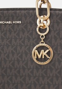 Borsa nera di pelle strutturata con hardware dorato, un charm circolare con il logo MK e il nome del brand "MICHAEL KORS" impresso sulla superficie.