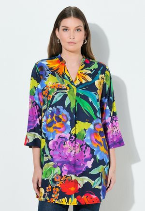 Vrouw draagt een loszittende, kleurrijke bloes met bloemenprint en driekwart mouwen, staat voor een witte muur en kijkt recht vooruit met een neutrale uitdrukking.