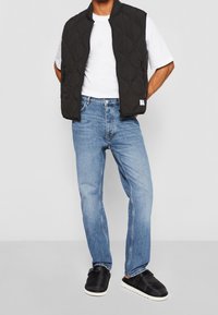 Chaleco acolchado negro sobre una camiseta blanca, combinado con jeans rectos de color azul claro y zapatos negros tipo slip-on con suelas blancas.