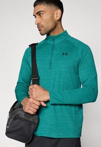 Τιρκουάζ αθλητική μπλούζα με φερμουάρ μέχρι τη μέση, υφασμάτινη με υφή, μακριά μανίκια και μαύρο λογότυπο. Το άτομο κρατά μια μαύρη γυμναστηριακή τσάντα με τσέπη από δίχτυ.