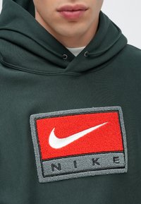 Σκούρο πράσινο φούτερ με κουκούλα που διαθέτει κόκκινο και γκρι υφασμάτινο λογότυπο της Nike με λευκό σβήσιμο. Κουκούλα με κορδόνια, απαλό ύφασμα και ελάχιστη ραφή.
