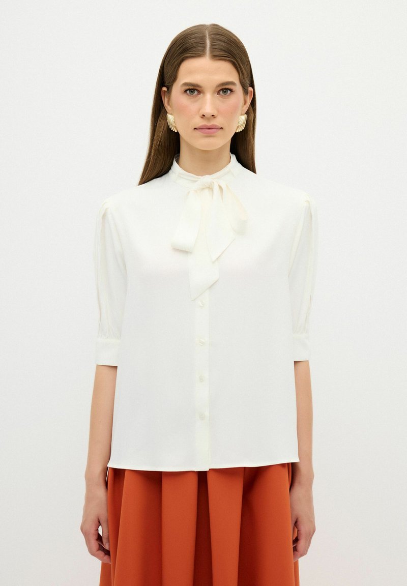 Blusa bianca con colletto alto e cravatta, chiusura a bottoni frontale, maniche corte a palloncino e superficie liscia. Indossata con una gonna color ruggine.