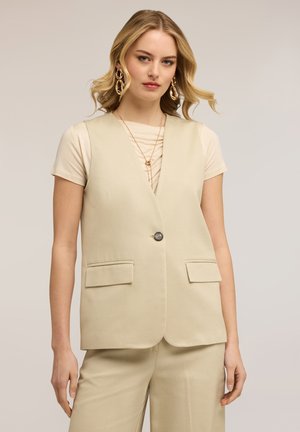 Blondynka ubrana w beżową kamizelkę na guziki bez rękawów i dopasowane spodnie, z złotymi geometrycznymi kolczykami i warstwowymi naszyjnikami.