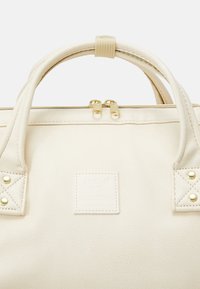 Sac beige en matériau synthétique texturé avec deux poignées, fermeture zippée et une étiquette rectangulaire portant le logo et la marque. Accents en métal doré.