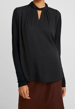 Vrouw draagt een zwarte blouse met lange mouwen, een gedraaide kraag en keyhole-halslijn, gecombineerd met een gestructureerde bruine rok.