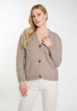 Blonde woman wearing a beige knit buttoned cardigan and white pants, posing with one hand near her chest and the other by her side.