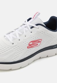 White athletic shoe with breathable mesh upper, red logo accent, navy details, and thick cushioned sole. Features lace-up design and textured surface.