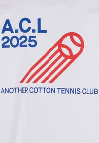 Balts kokvilnas T-krekls ar zilo tekstu "A.C.L 2025" un sarkano grafiku ar tenisa bumbu ar līnijām, ar uzrakstu "CITS KOKVILNAS TENISA KLUBS."