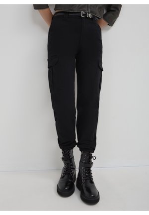 Personne portant un pantalon cargo noir avec poches latérales, ceinture noire et bottes de combat noires brillantes à lacets, debout sur un fond blanc.