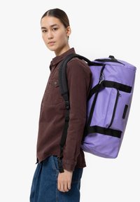 Zaino viola con accenti neri, tessuto resistente e tasca laterale. Indossato sulla schiena, abbinato a una camicia button-up marrone e jeans blu.