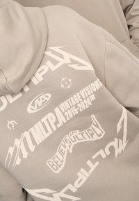 Sweat à capuche gris avec de grands graphismes imprimés blancs incluant « MULTIPLY » et « CROIRE EN MULTIPLY », avec divers logos et motifs partout.