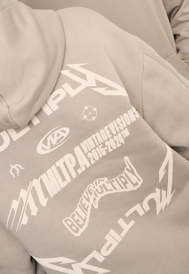 Sweat à capuche gris avec de grands graphismes imprimés blancs incluant « MULTIPLY » et « CROIRE EN MULTIPLY », avec divers logos et motifs partout.