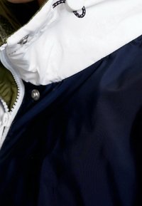 Laivastonsininen ja valkoinen eristetty takki, jossa on vetoketjusuljenta, napit ja nyörikiinnitys hupussa. Ulkopinta on sileä ja kiiltävä.