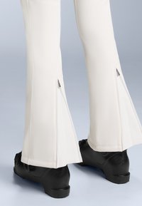 Pantalonii albi cu evazare au fermoare pe partea de jos a piciorului, asortați cu încălțăminte neagră. Materialul este neted, cu un ușor luciu.