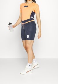Top sportivo a maniche corte arancione e blu scuro, con un taglio a chiave. Abbinato a pantaloni corti blu scuro, con un cordoncino rosa, e una bottiglia d'acqua.