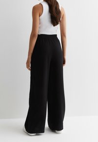 Zwarte, wijdvallende broek van een gestructureerde stof, met een elastische tailleband en een losse pasvorm, gecombineerd met een witte mouwloze top en witte schoenen.