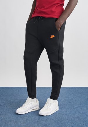 Osoba ubrana w czarne spodnie dresowe Nike z pomarańczowym logo, czerwoną koszulkę oraz białe sneakersy Nike Air Max, stojąca na niebieskim dywanie.