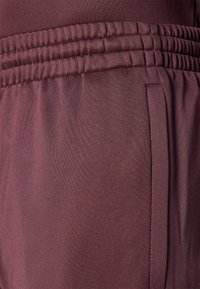 Tecido castanho com uma textura suave, apresentando uma faixa elástica na cintura e um bolso lateral. O close-up mostra os detalhes da costura.