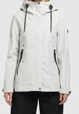 Chaqueta blanca con capucha, bolsillos con cremalleras negras, cordones ajustables y puños con botones, usada sobre pantalones negros.