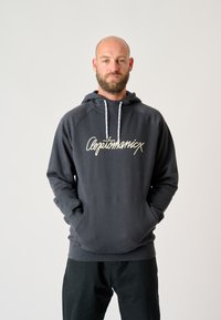 Sudadera gris oscuro con un bolsillo frontal, cordones blancos y un gran logo de script de color claro en el pecho. Material de algodón suave.