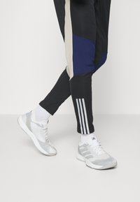 Sportliche Schuhe in Weiß mit silbernen Akzenten, ausgestattet mit einer strukturierten Oberfläche und Schnürsenkeln. Schwarze und blaue Jogginghosen mit drei weißen Streifen am Bein.