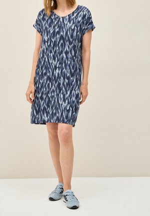 Woman wearing a knee-length blue patterned dress and light blue sneakers standing against a beige background.