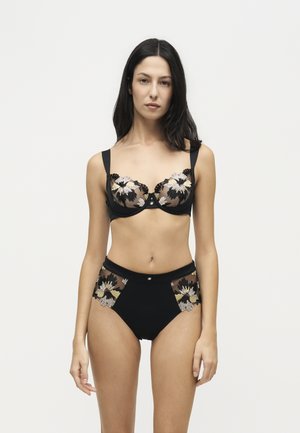 Femme aux longs cheveux foncés portant un soutien-gorge noir et une culotte taille haute avec une broderie florale transparente, debout devant un fond clair uni.