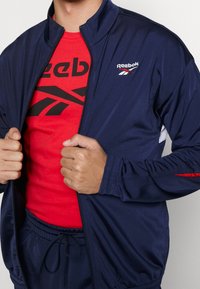 Tmavě modrá bunda na zip s červen ými doplňky, na ní je logo Reebok. Pod ní je červené tričko s černým grafikou Reebok. Viditelný elastický pas.