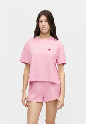 Vrouw die een loszittend roze shirt met korte mouwen en bijpassende korte broek draagt, met een klein rood hartje op de linkerborst van het shirt.