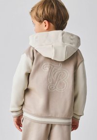 Bluza z kapturem w kolorze beżowym i kremowym, z haftowanym napisem "BAKE" na kapturze oraz stylizowanym napisem "TB" na plecach. Miękka w dotyku, z prążkowanymi mankietami.