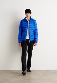 Blå pufferjakke med et kviltet design, båret over en hvid skjorte, parret med sorte bukser og en sort beanie. Står foran en neutral baggrund.