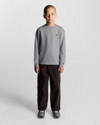 Sweatshirt gris clair avec un petit logo jaune, associé à un pantalon noir et des baskets grises, sur une personne se tenant contre un fond uni.