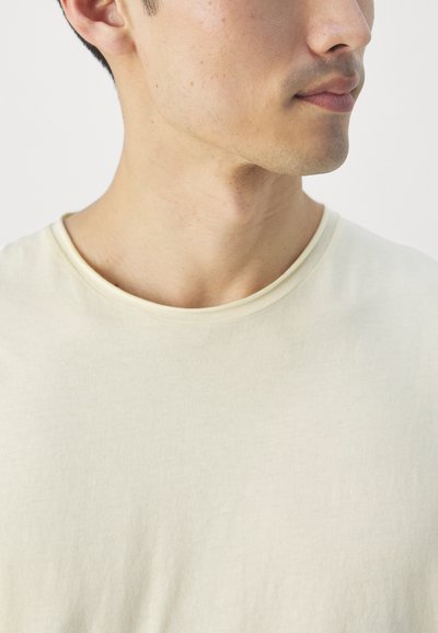 T-shirt en coton beige clair avec un col rond. Le tissu a une apparence lisse, légèrement texturée et une coupe décontractée.