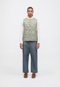 Άνδρας που στέκεται με το πρόσωπο μπροστά, φορώντας ένα ανοιχτό πράσινο γαζωμένο γιλέκο, λευκό πουκάμισο, φαρδιά μπλε τζιν και καφέ δερμάτινα παπούτσια με κορδόνια.