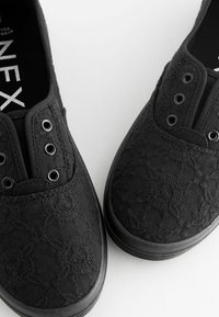 Schwarze Stoff-Slipper mit strukturiertem Blumenmuster, runder Zehenpartie und silbernen Ösen. Weiche schwarze Innenausstattung und flexible Gummisohle.