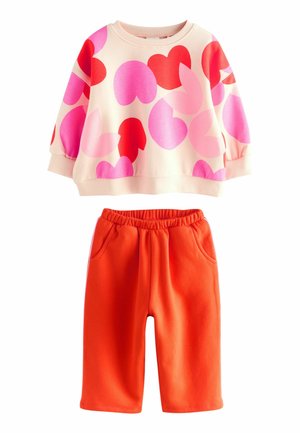 Crème sweatshirt met lange mouwen met rode en roze hartvormen, gecombineerd met felrode elastische broek met zijzakken.