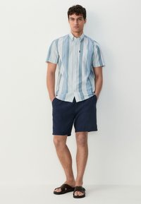 Srajca s kratkimi rokavi, svetlo modre barve z žičničnimi črtami, s kragnom na gumb, v kombinaciji z mornarskimi kratkimi hlačami in temno rjavimi natikači. Podrobnost žepa na srajci.