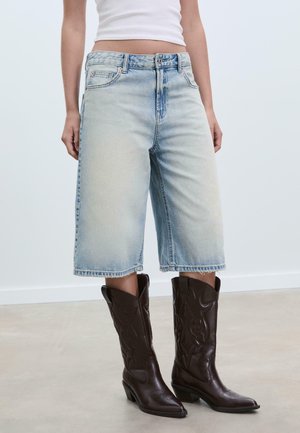 Shorts en denim bleu clair à taille haute avec un ourlet effiloché, associés à des bottes de cowboy en cuir marron foncé avec des coutures délicates et un bout pointu.