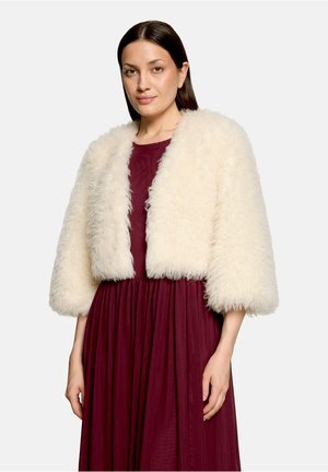 Femme aux cheveux raides et foncés porte une veste courte blanche et duveteuse sur une longue robe bordeaux, sur un fond clair uni.