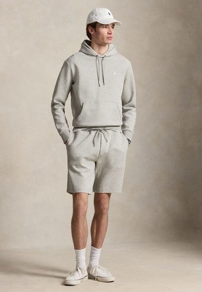 Jeune homme portant un sweat à capuche gris clair et un short assorti, casquette blanche, chaussettes blanches et baskets blanches, debout les mains dans les poches.