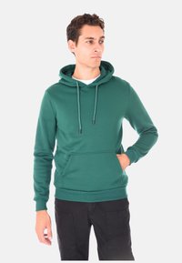 Uomo con felpa verde con cappuccio e tasca frontale, pantaloni neri, che guarda di lato con una mano in tasca, in piedi contro uno sfondo neutro.