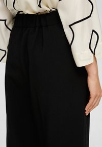 Zwarte high-waisted pantalon met een gladde textuur, voorzien van een geplooide tailleband. Een crèmekleurige blouse met abstracte zwarte patronen die gedeeltelijk zichtbaar zijn.