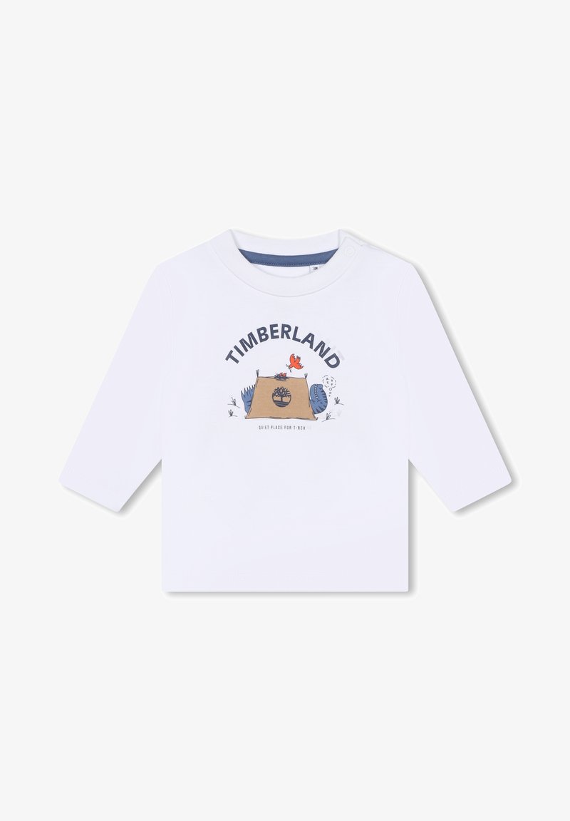 T-shirt bianca a maniche lunghe con stampa grafica "TIMBERLAND", una scena di campeggio e elementi cartooneschi colorati. Chiusura a scatto sulla spalla.