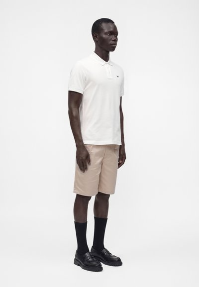 Homme debout portant un polo blanc à manches courtes, un short beige au genou, des chaussettes noires et des mocassins noirs sur un fond blanc uni.