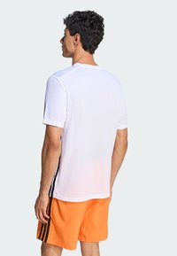 Camiseta deportiva blanca de manga corta con franjas negras en los laterales, combinada con pantalones cortos naranja. La tela parece ligera y suave.