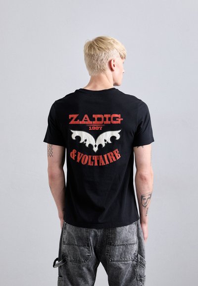 Blond young man with tattoos wears a black Zadig & Voltaire shirt with red text and white graphic on the back, paired with gray cargo pants.