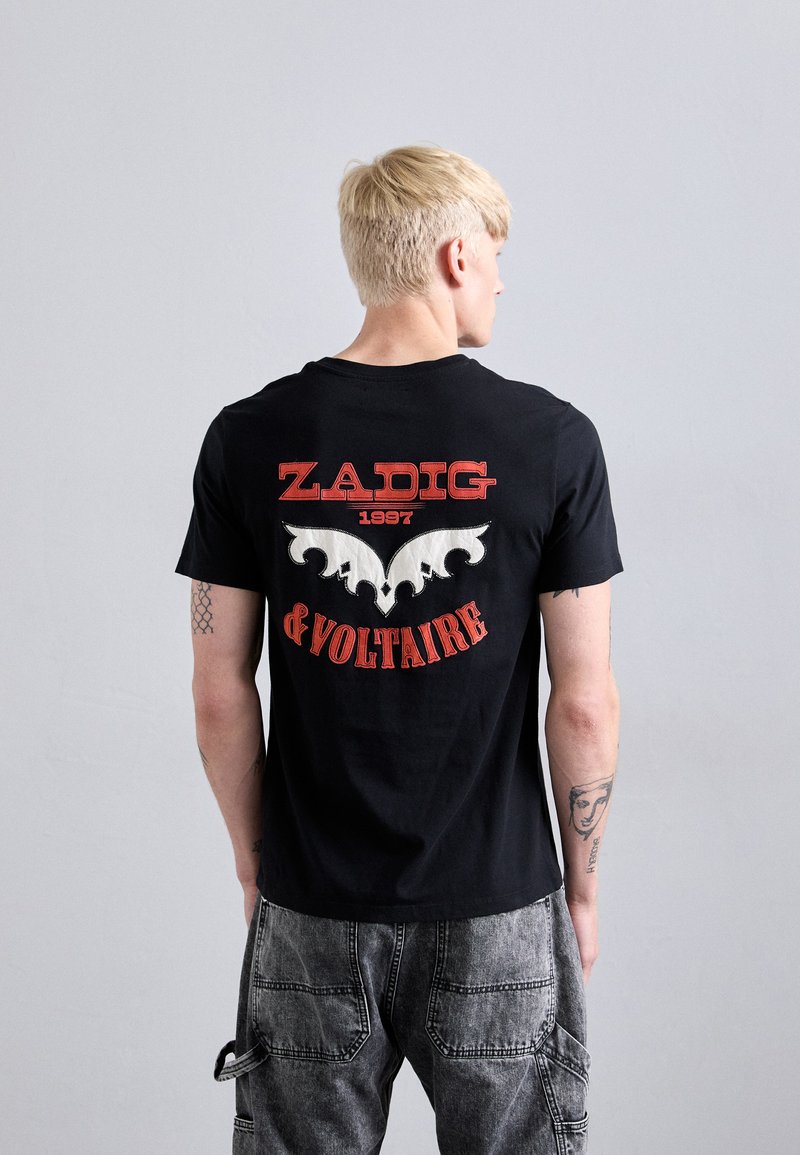 Jeune homme blond avec des tatouages porte une chemise noire Zadig & Voltaire avec un texte rouge et un graphique blanc au dos, associée à un pantalon cargo gris.