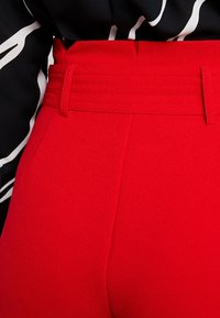 Žena v červených kalhotách s vysokým pasem, širokým páskem a poutky na opasek, v kombinaci s černobílým vzorovaným topem.