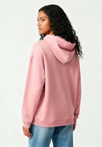 Felpa con cappuccio rosa, dotata di polsini e bordo a coste. Il tessuto sembra morbido, presenta una vestibilità ampia e un design semplice. Lo sfondo è neutro.