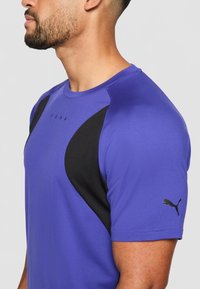 Blå trenings t-skjorte med svarte detaljer på skuldrene, PUMA-logo på brystet og ermet, rund hals, korte ermer og glatt tekstur.